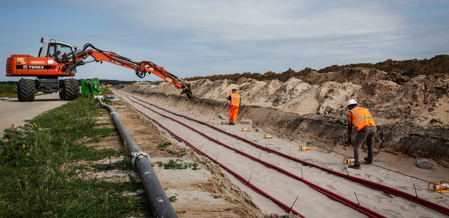 Aangeleverde projectfoto: een open sleuf met kabels, een graafmachine en medewerkers aan het werk.